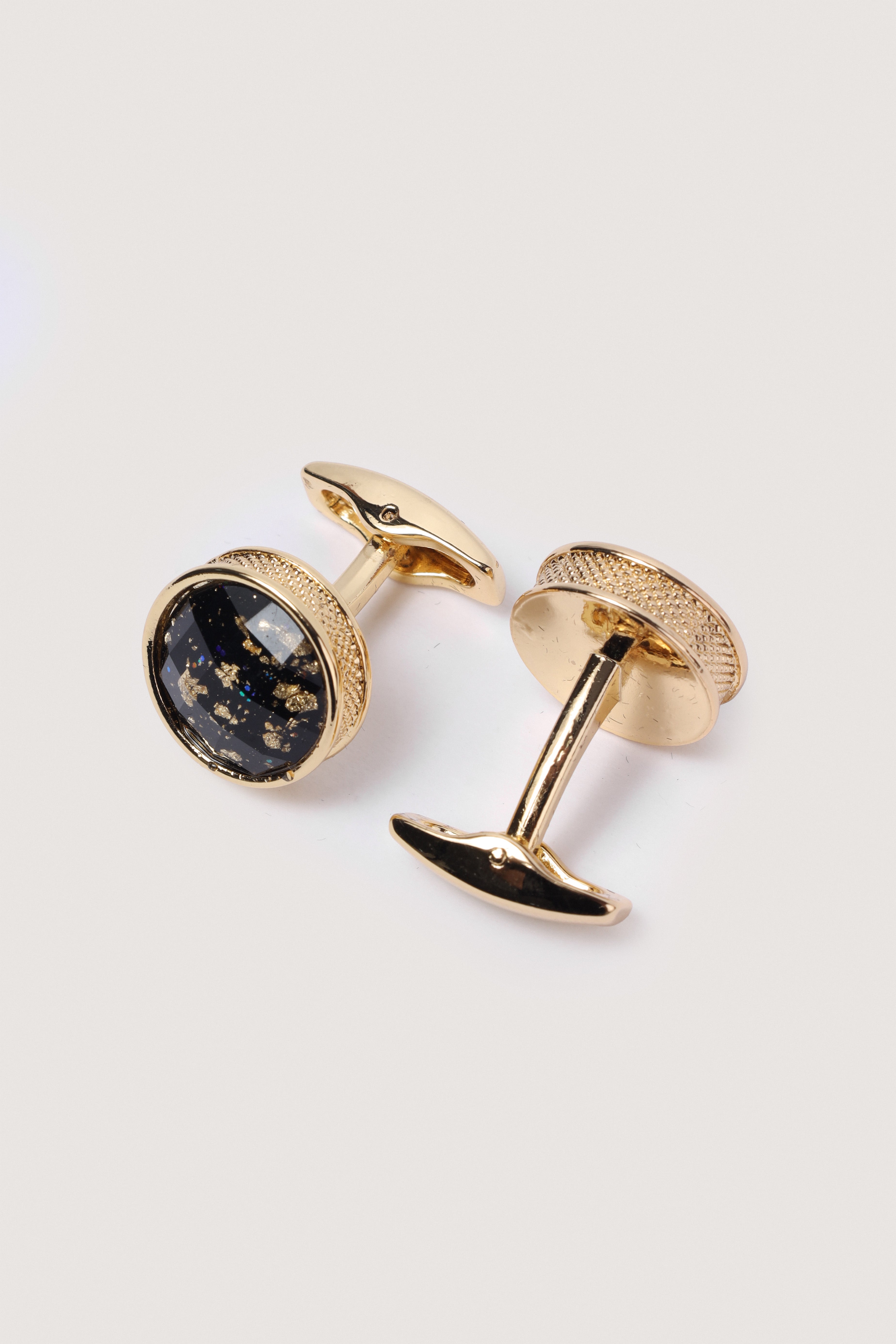 A pair of gold cufflinks with round black enamel faces containing tiny gold flecks, shown on a light gray background.