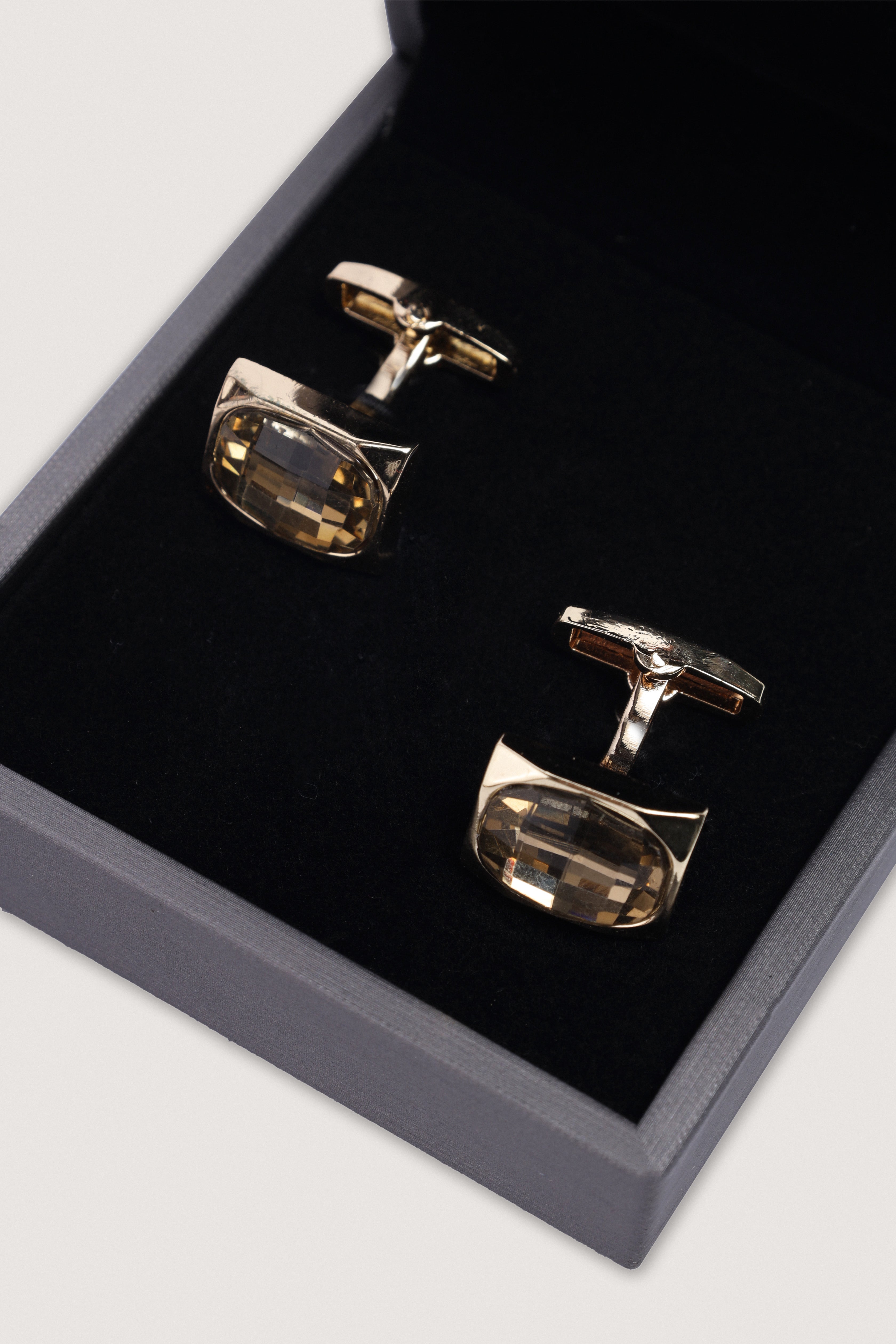 A pair of men's cufflinks with polished gold-toned metal and faceted clear crystal stones, presented inside a dark gray box with a black velvet lining.